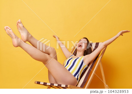 Indoor shot of overjoyed Caucasian young woman wearing striped one-piece swimsuit posing on wooden chair isolated on yellow background, rejoicing vacation, spreading arms 104328953