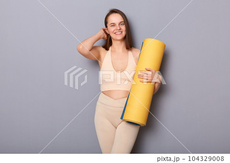 Image of pretty cute charming woman wearing beige top and leggins holding fitness mat isolated over gray background, touching her hair, smiling at camera, visiting gym for improving body shape Image of pretty cute charming woman wearing beige top and leggins holding fitness mat isolated over gray background, touching her hair, smiling at camera, visiting gym for improving body shape 104329008