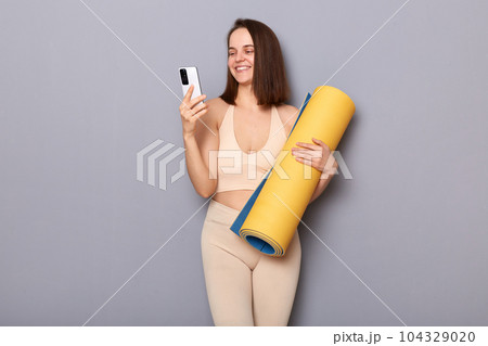 Indoor shot of smiling delighted sporty woman wearing sportswear with fitness mat in hands isolated over gray background, using her smart phone, checking burning calories in app Indoor shot of smiling delighted sporty woman wearing sportswear with fitness mat in hands isolated over gray background, using her smart phone, checking burning calories in app 104329020