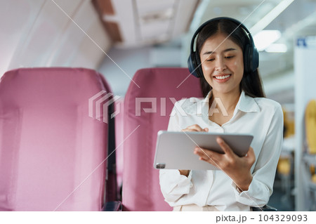 Young Asian attractive business woman passenger sitting on business class luxury plane while wearing wireless headphone for relax and working with tablet during the flight. Young Asian attractive business woman passenger sitting on business class luxury plane while wearing wireless headphone for relax and working with tablet during the flight. 104329093