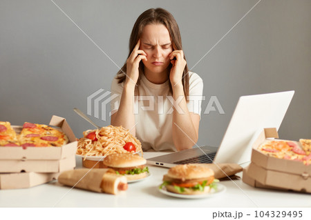 Indoor shot of sick unhealthy woman sitting in front of laptop among junk food, having terrible headache, being tire and exhausted, isolated over gray background. 104329495