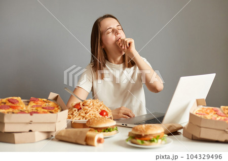 Image of sleepy tired woman working or watching movie on laptop during long hours isolated over gray background, being surrounded with pizza, pasta, hotdogs, hamburgers. 104329496