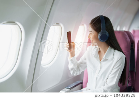 Young Asian attractive business woman passenger sitting on business class luxury plane while wearing wireless headphone and using smart phone mobile for relax during the flight. 104329529