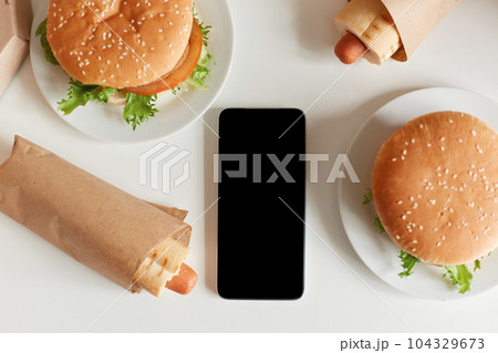 Top view image of junk food on table, tasty sausage in dough and big hamburgers, smart phone with black blank screen, copy space for advertisement or promotional text. 104329673