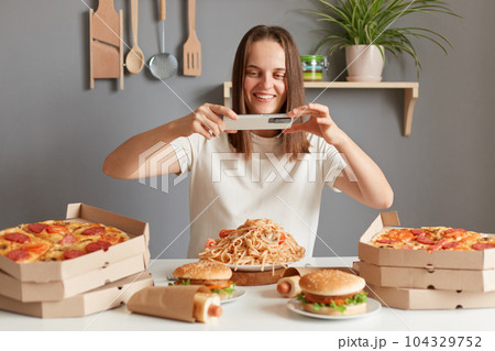 Image of smiling attractive woman food blogger with brown hair wearing white T-shirt sitting at table, taking photo of her dinner for her vlog, wants to tell about fast food. Image of smiling attractive woman food blogger with brown hair wearing white T-shirt sitting at table, taking photo of her dinner for her vlog, wants to tell about fast food. 104329752