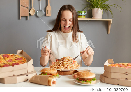 Portrait if amazed excited extremely hungry woman wearing white T-shirt sitting at table in kitchen, delivering lots jink food dishes, eating noodles, expressing happiness. 104329760