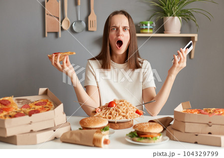 Indoor shot of shocked irritated woman with brown hair wearing white t shirt sitting in kitchen at table and holding smart phone and slice pizza, looking at camera with shocked face. Indoor shot of shocked irritated woman with brown hair wearing white t shirt sitting in kitchen at table and holding smart phone and slice pizza, looking at camera with shocked face. 104329799