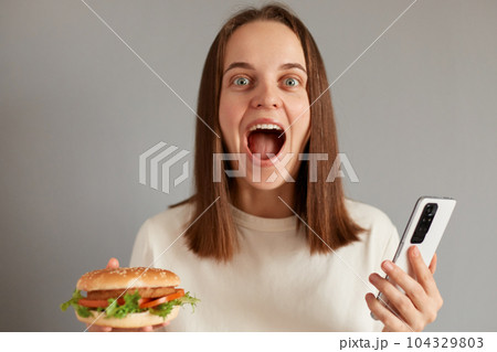 Portrait of amazed young adult caucasian woman wearing white t-shirt holding american classic fast food burger, using mobile phone isolated over gray background, screaming with excitement. Portrait of amazed young adult caucasian woman wearing white t-shirt holding american classic fast food burger, using mobile phone isolated over gray background, screaming with excitement. 104329803