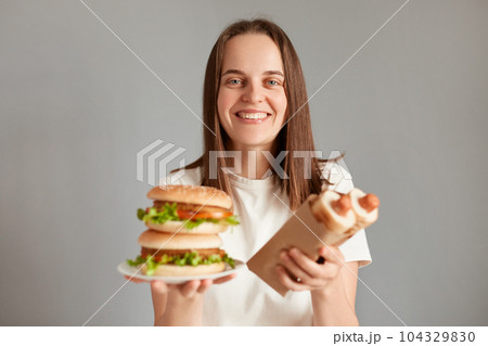 Horizontal shot of smiling satisfied female with brown hair, standing isolated on gray background, holding hot dogs and american hamburger, enjoying fast food. 104329830