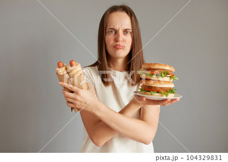 Portrait of frustrated woman holding hot dog and big sandwich, wearing white T-shirt posing isolated over gray background, keeps hands crossed, thinks eating fast food is very harmful for your health. Portrait of frustrated woman holding hot dog and big sandwich, wearing white T-shirt posing isolated over gray background, keeps hands crossed, thinks eating fast food is very harmful for your health. 104329831