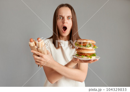 Indoor shot of shocked scared woman looking at camera with big eyes and open mouth, keeps arms crossed, holding sandwich and hot dogs, keeps diet, expressing astonishment. Indoor shot of shocked scared woman looking at camera with big eyes and open mouth, keeps arms crossed, holding sandwich and hot dogs, keeps diet, expressing astonishment. 104329833