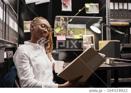 Policewoman reading investigation report while drinking coffee in office. African american woman police detective holding crime case file folder and analyzing evidence at night Policewoman reading investigation report while drinking coffee in office. African american woman police detective holding crime case file folder and analyzing evidence at night 104329922