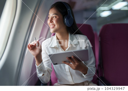 Young Asian attractive business woman passenger sitting on business class luxury plane while wearing wireless headphone for relax and working with tablet during the flight. Young Asian attractive business woman passenger sitting on business class luxury plane while wearing wireless headphone for relax and working with tablet during the flight. 104329947