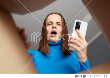 Astonished Caucasian young adult woman wearing blue turtleneck opening carton box, looking inside package with shocked face, holding smart phone in hand, getting wrong parcel, view from bottom. 104330043