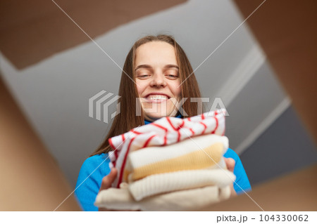 Attractive optimistic woman customer wearing blue turtleneck packing her clothing into cardboard box, relocating to her new flat, view from the bottom of box. 104330062
