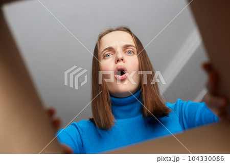 Shocked disappointed woman customer wearing blue turtleneck opening cardboard box with broken or wrong order, looking inside parcel with widely open mouth and big eyes. Shocked disappointed woman customer wearing blue turtleneck opening cardboard box with broken or wrong order, looking inside parcel with widely open mouth and big eyes. 104330086