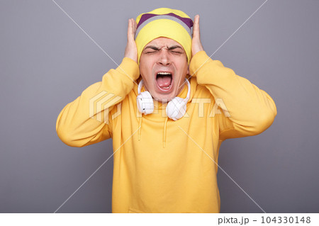 Portrait of shocked annoyed man wearing yellow hoodie, beanie hat and headphones, covering ears with hands, screaming, hearing loud noise, standing isolated over gray background. Portrait of shocked annoyed man wearing yellow hoodie, beanie hat and headphones, covering ears with hands, screaming, hearing loud noise, standing isolated over gray background. 104330148
