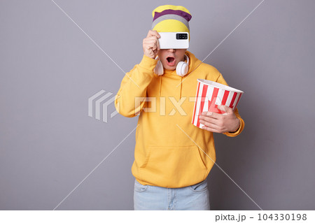 Photo of shocked surprised unknown man wearing yellow hoodie and beanie hat, posing in cinema against gray wall, holding paper bucket with popcorn, covering his eyes with smart phone, hiding. Photo of shocked surprised unknown man wearing yellow hoodie and beanie hat, posing in cinema against gray wall, holding paper bucket with popcorn, covering his eyes with smart phone, hiding. 104330198