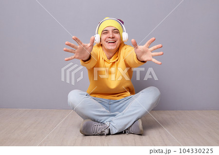 Come into my arms. Portrait of smiling satisfied man wearing beanie hat, yellow hoodie and jeans posing against gray wall, outstretching hands, wants to embrace, being happy. 104330225