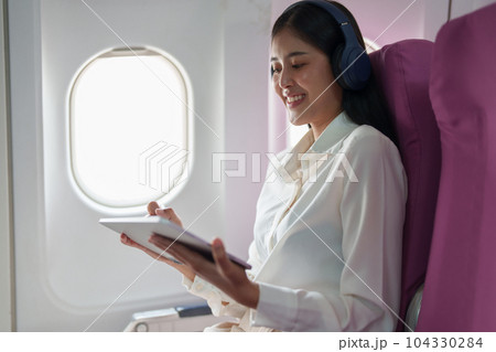 Young Asian attractive business woman passenger sitting on business class luxury plane while wearing wireless headphone for relax and working with tablet during the flight. 104330284