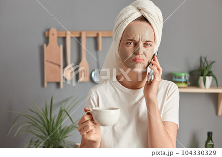 Sad upset woman with cosmetic mask drinking coffee or tea, talking on cell phone in kitchen, offended female wrapped in towel answering call by phone during beauty procedures, hearing unpleasant news. 104330329