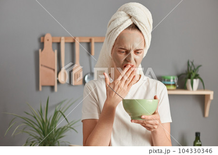 Upset Caucasian woman wearing white t-shirt and wrapped towel on her hair doing cosmetic procedures and having breakfast, covering mouth with palm, realized that the food is spoiled 104330346