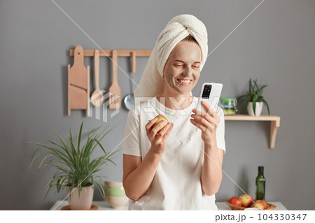 Indoor shot of smiling woman with treatment facial mask enjoying spa procedures at home, posing in the kitchen, eating apple and using cell phone, checking social networks. Indoor shot of smiling woman with treatment facial mask enjoying spa procedures at home, posing in the kitchen, eating apple and using cell phone, checking social networks. 104330347