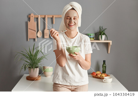 Indoor shot of Caucasian young adult woman wrapped white towel and dressed in T-shirt, eating her breakfast and doing skin care procedures, standing in kitchen at home. 104330419