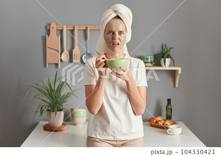 Photo of scared Caucasian woman with towel on head standing speechless indoor with beauty facial mask to reduce fine lines eats her breakfast, awakes early in morning, posing in kitchen. 104330421