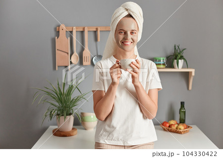 Indoor shot of young relaxed woman with cosmetic mask in bath towel on her head drinking hot beverage, tea or coffee in kitchen after taking shower bath at home. Beauty treatment, hydration. Indoor shot of young relaxed woman with cosmetic mask in bath towel on her head drinking hot beverage, tea or coffee in kitchen after taking shower bath at home. Beauty treatment, hydration. 104330422