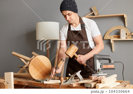 Portrait of young adult man carpenter wearing white t-shirt, black cap and brown apron, craftsman carve with a gouge in the hands on the workbench in carpentry. 104330664