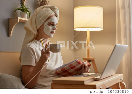Side view portrait of charming cute woman wearing white T-shirt and towel sitting on sofa at home, holding book or organizer, watching something on laptop, drinking coffee. Side view portrait of charming cute woman wearing white T-shirt and towel sitting on sofa at home, holding book or organizer, watching something on laptop, drinking coffee. 104330810