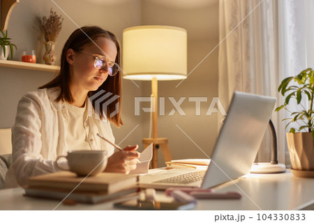 Indoor shot of focused woman wearing white shirt sitting on table and working on laptop near window, having concentrated facial expression, studying hard or works with a new project. Indoor shot of focused woman wearing white shirt sitting on table and working on laptop near window, having concentrated facial expression, studying hard or works with a new project. 104330833