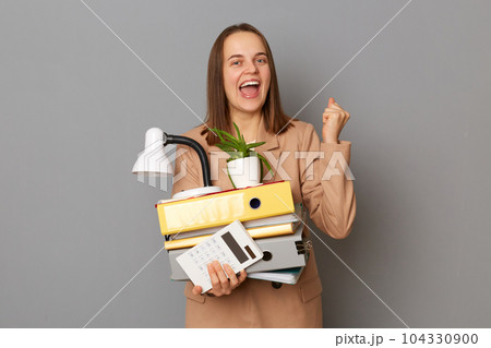 Happy young pretty woman wearing beige jacket holding documents and her office stuff posing isolated over gray background, successfully passed the interview and am ready to start working 104330900