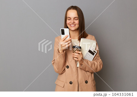 Image of attractive woman with brown hair wearing beige jacket standing isolated over gray background, holding papers documents and takeaway coffee, using mobile phone, smiling. 104331061