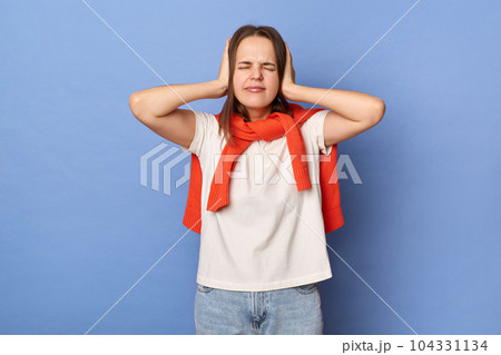 Portrait of despair dark haired woman wearing white T-shirt and orange sweater tied over shoulders, covering ears with hands, avoids noise, posing isolated on blue background. Portrait of despair dark haired woman wearing white T-shirt and orange sweater tied over shoulders, covering ears with hands, avoids noise, posing isolated on blue background. 104331134
