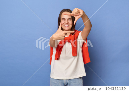 Portrait of cheerful optimistic woman photographer wearing casual style clothing standing isolated over blue background, looking at camera through frame from fingers, focus. 104331139