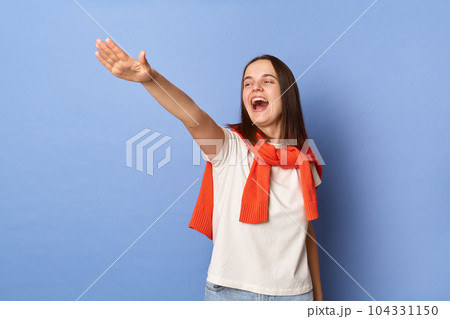 Photo of excited happy smile young adult woman dressed white T-shirt and orange jumper tied over shoulders standing against blue wall, standing with raised arms, screaming. 104331150