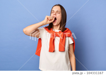 Indoor shot of sleepless tired exhausted woman dressed white T-shirt and tied sweater over shoulders standing isolated on blue background, yawning, covering mouth with fist, needs to have nap. 104331167