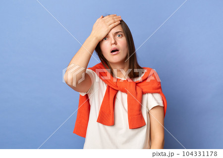 Portrait of shocked disappointed brown haired woman wearing T-shirt and orange sweater tied over shoulders, showing facepalm gesture, loosing, having problems, standing isolated on blue background. 104331178