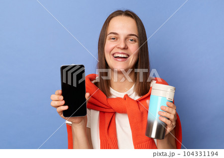 Portrait of cute smiling woman in white T-shirt and orange sweater tied over shoulders, showing empty display of her phone, holding thermos with coffee or tea, standing isolated on blue background. Portrait of cute smiling woman in white T-shirt and orange sweater tied over shoulders, showing empty display of her phone, holding thermos with coffee or tea, standing isolated on blue background. 104331194