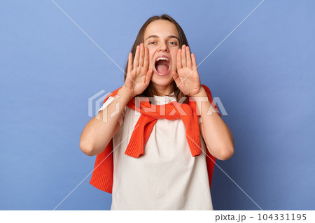 Indoor shot of beautiful attractive woman wearing white T-shirt and orange sweater tied over shoulders standing isolated on blue background, screaming loud with hands near mouth, making announcement. 104331195