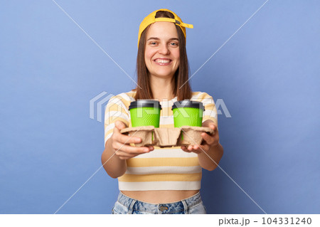 Portrait of positive teenager girl wearing striped T-shirt and baseball cap posing isolated over blue background, female working as courier, delivering takeaway coffee. Portrait of positive teenager girl wearing striped T-shirt and baseball cap posing isolated over blue background, female working as courier, delivering takeaway coffee. 104331240
