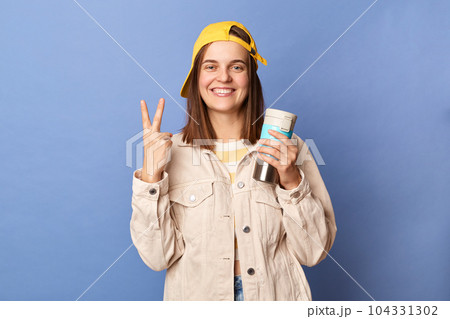 Photo of smiling delighted brown haired teenager girl wearing baseball cap and jacket, drinking coffee from thermos, showing victory gesture, v sign, posing isolated over blue background. Photo of smiling delighted brown haired teenager girl wearing baseball cap and jacket, drinking coffee from thermos, showing victory gesture, v sign, posing isolated over blue background. 104331302