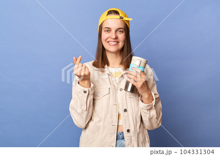 Indoor shot of delighted satisfied teenager girl wearing baseball cap and jacket posing isolated over blue background, showing mini love gesture, drinking hot beverage from thermos. 104331304