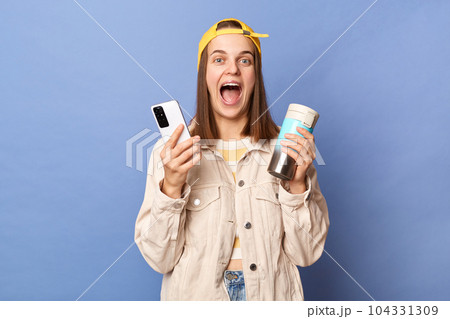 Photo of excited amazed positive brown haired teenager girl wearing baseball cap and jacket posing isolated over blue background, holding thermos, using cell phone, reading message with good news. Photo of excited amazed positive brown haired teenager girl wearing baseball cap and jacket posing isolated over blue background, holding thermos, using cell phone, reading message with good news. 104331309