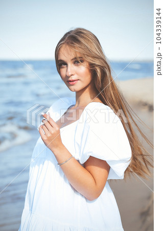 Happy, beautiful woman on the ocean beach standing in a white summer dress 104339144