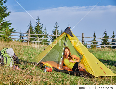 Woman hiker camping in mountains. Young lady traveling, hiking in hills in summer. Pretty, slim woman sitting in tent in campsite, holding flower, admiring. Concept of harmony with nature. 104339279