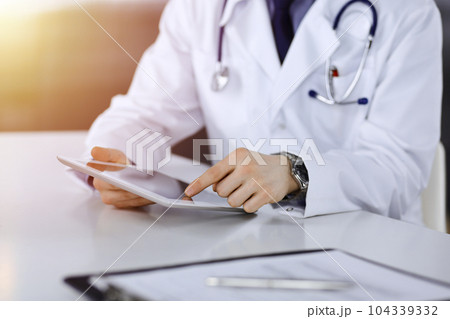 Unknown male doctor sitting and working with tablet computer iin a darkened clinic, glare of light on the background, close-up of hands 104339332