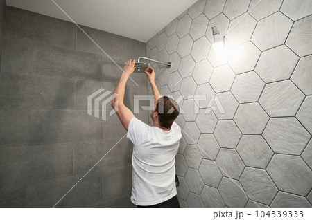 Male worker fixing bathroom showerhead while working on bathroom renovation. Back view of man plumber installing high pressure bath showerhead at home. 104339333